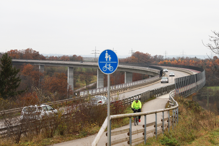 Es ist die Brücke über dem Viadukt zu sehen. Vor der Brücke steht ein blaues rundes Schild. Auf dem Schild ist oben eine Person mit einem Kind an der Hand abgezeichnet. Darunter ist ein Strich. Darunter ist ein Fahrrad abgezeichnet. 
