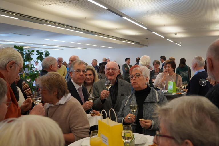 Besucherinnen und Besucher stehen im Raum verteilt und unterhalten sich bei Gebäck und Getränken.